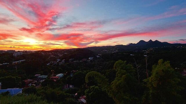 Ciel de feu rose et orange au-dessus de la ville. Le soleil couchant cr&eacute;e un contraste saisissant avec la silhouette urbaine et la v&eacute;g&eacute;tation luxuriante. Cr&eacute;puscule d'une intensit&eacute; sublime.