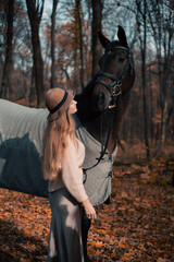 Woman stands beside horse in forest during fall season while wearing a hat and sweater © Тамара Печеная