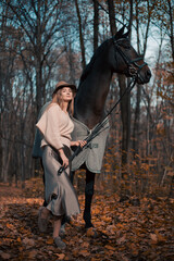 Naklejka premium Woman stands beside a horse in a forest with autumn leaves during daylight in October
