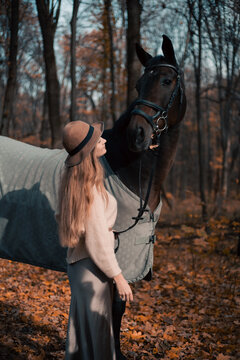Woman stands beside horse in forest during fall season while wearing a hat and sweater