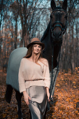 Naklejka premium Woman stands beside horse in forest during fall season while wearing a hat and sweater