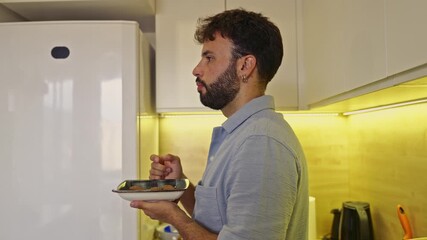 A bearded young man takes a moment to savor a small, tasty snack in his bright, modern kitchen. The video features clear chewing sounds, capturing an authentic moment of enjoyment and daily routine.
