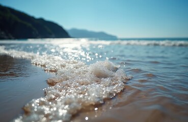 Close-up of ocean waves rolling onto sandy beach. Sunlight reflects on wet sand, foam. Blue sky provides serene backdrop to this coastal scene. Tranquil water meets shore creating peaceful atmosphere.