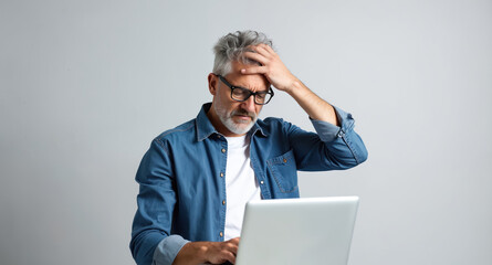 Mature man with glasses feels stressed or shocked while reading email on laptop computer. He rubs his forehead in despair over digital information on gray background.