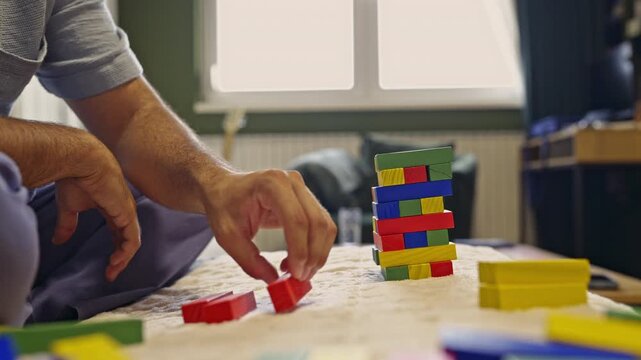 An adult's hands carefully stack colorful wooden blocks to build a tower, which eventually collapses, capturing the fun and challenge of the game with accompanying sounds.
