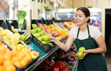 Young woman seller in apron lays out apples in supermarket