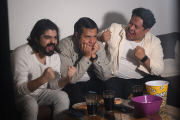 Two friends cheering while the third shows anxiety during broadcast, watching tv