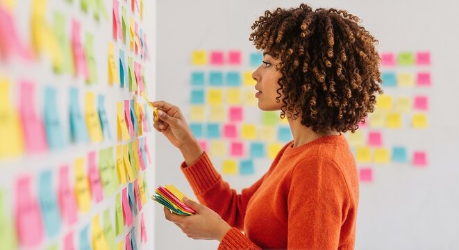 Woman with curly hair placing sticky notes on a wall covered with colorful sticky notes for planning