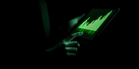 Person using tablet device in dark environment with green data visualization on screen, creating dramatic cybersecurity atmosphere.