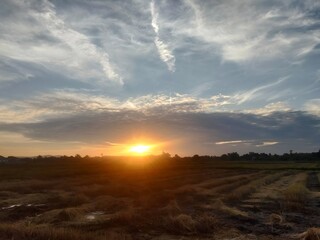 The image depicts a rural landscape at sunset, with the sun partly below the horizon casting a warm, golden glow across the scene. The sky is filled with a mix of wispy and scattered clouds