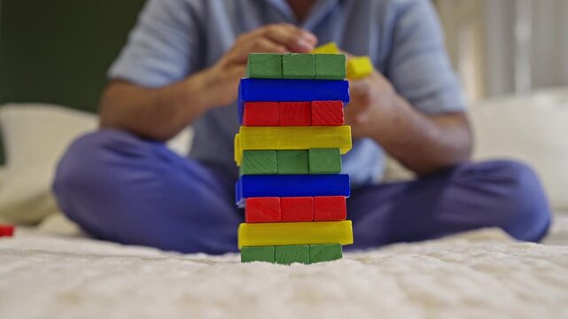 A man in pajamas is focused on a challenging Jenga-style block game, carefully removing and stacking colorful wooden blocks. The tower wobbles precariously before eventually collapsing, with sounds.