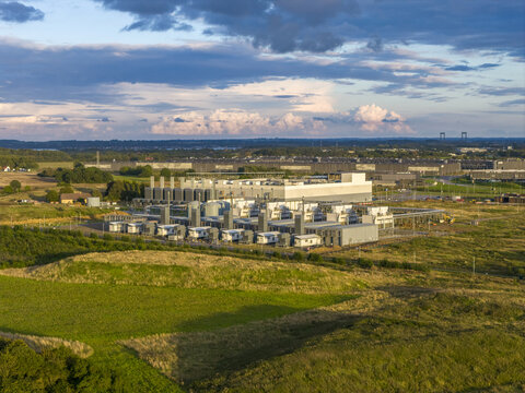 Aerial view of a modern industrial complex bathed in the warm glow of the setting sun amid lush green fields under a dramatic sky, Fredericia, Southern Denmark, Denmark.