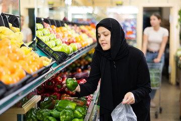 Asian girl in hijab buys ripe bell peppers in the supermarket