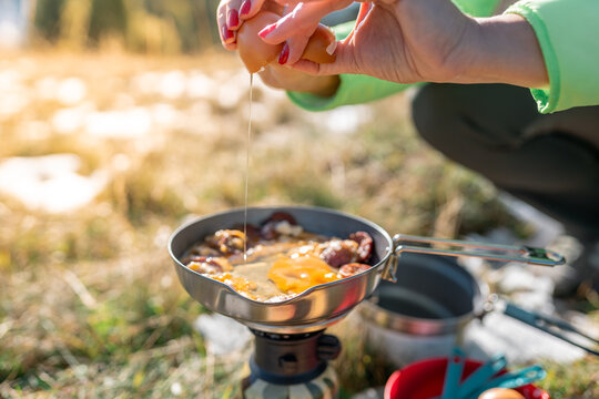 Mountain picnic scene with a woman preparing a simple outdoor meal using eggs, tomatoes, and bread beside a portable camping stove in warm morning light.