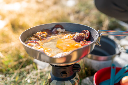 Mountain picnic scene with a woman preparing a simple outdoor meal using eggs, tomatoes, and bread beside a portable camping stove in warm morning light. - Powered by Adobe