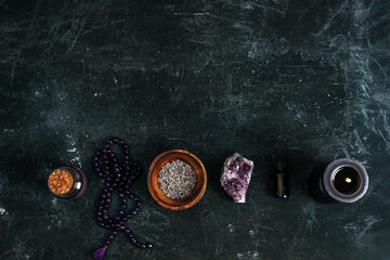 Lavender dried flowers in a bowl, amethyst crystal, beads, oils and candle on dark textured background. Magical items, alternative healing, esoteric ritual concept. Top view, flat lay