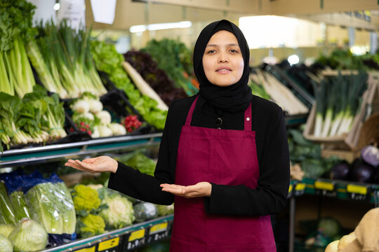 Fototapeta Female seller in a hijab and an apron offers to buy vegetables and fruits in the grocery department of a supermarket