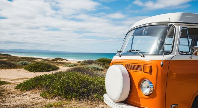 Classic orange and white vintage volkswagen t2 camper van parked on a sandy beach near the ocean under a bright blue sky with scattered clouds, suggesting a summer road trip or vacation