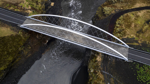 Aerial view of a sleek, modern bridge gracefully spanning the dark, swirling waters, a stark contrast against the rugged, moss-covered banks, Hvolsvollur, Rangarbing eystra, Iceland.