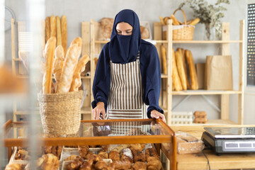 Shopping area, showcase, counter in bakery. Muslim girl seller in hijab employee fills window and...