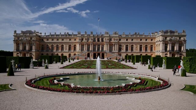 A wide-angle video shot of a grand palace with ornate gardens and a central fountain, showcasing classic architecture under a clear blue sky.