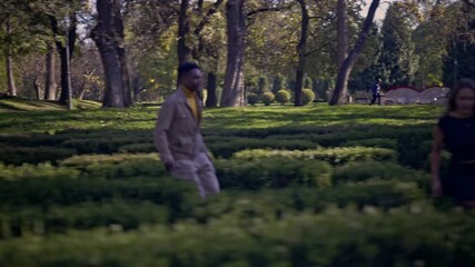 A joyful young couple shares a playful and romantic chase through the beautifully manicured hedges of a green park maze. They laugh and connect, embracing moments of happiness and freedom outdoors.