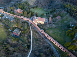 Aerial view of the Portico di San Luca stretching through the verdant hills, a terracotta ribbon connecting urbanity to spiritual heights, Bologna, Emilia-Romagna, Italy.
