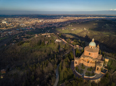 Aerial view of the Sanctuary of San Luca standing proudly amidst the rolling green hills, its terracotta facade glowing warmly against the cool shadows, Bologna, Emilia-Romagna, Italy.