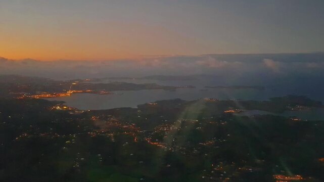 Vue a&eacute;rienne de nuit. Le cr&eacute;puscule enveloppe une baie bord&eacute;e de lumi&egrave;res. Le paysage s'&eacute;tale, mariant le ciel orang&eacute; et l'obscurit&eacute; de l'eau. Une atmosph&egrave;re sublime.