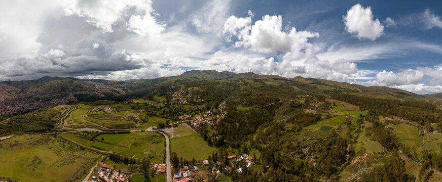 Panoramic view of the rural countryside surrounding Sacsayhuaman ruins in Cusco, Peru. Features green fields, eucalyptus forests, and a glimpse of the city under a dramatic cloudy sky.