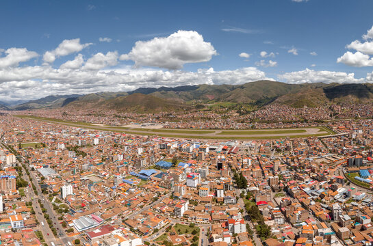 Wide aerial panorama of modern Cusco, Peru. Features the busy Avenida de la Cultura highway running parallel to the Alejandro Velasco Astete Airport runway amidst the urban valley.