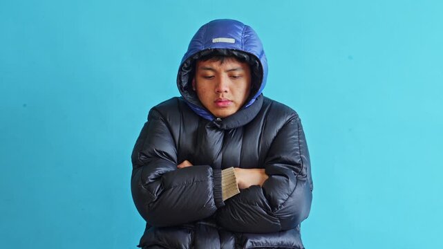 Young asian man shivering in puffer jacket feeling cold and uncomfortable isolated on blue background