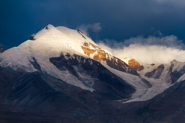 Telephoto view of golden alpenglow illuminating the glaciated summit of Muztagh Ata against stormy...