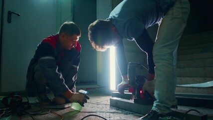 two caucasian men fixing basement light, crouched beside bright vertical tube lamp, tools and cables scattered on concrete floor, electrician guides assistant through wiring, hands