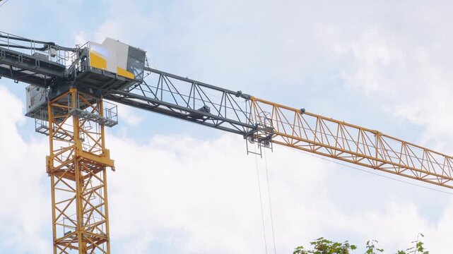 Yellow and grey industrial tower crane rotating at a construction site. The massive machinery moves slowly against a cloudy blue sky