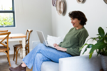 Woman sitting on grey sofa using silver laptop near potted plant in living room, copy space