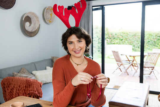 Woman sitting at table in living room crafting holiday ribbon wearing red antler headband