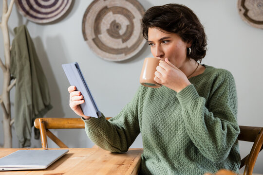 woman in late twenties using tablet and sipping mug at kitchen nook with laptop and baskets - Powered by Adobe