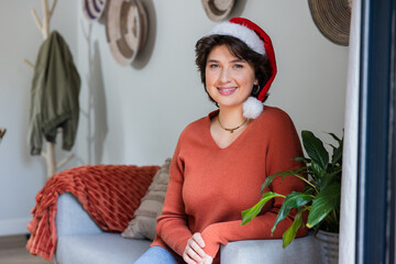 Woman sitting on light gray sofa wearing Santa hat and knit sweater amid decorative woven hats