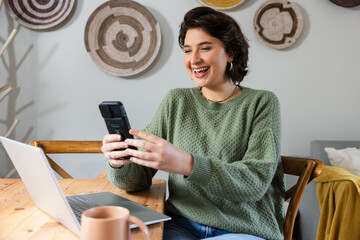 Woman wearing green sweater sitting at dining table holding smartphone with laptop and coffee mug