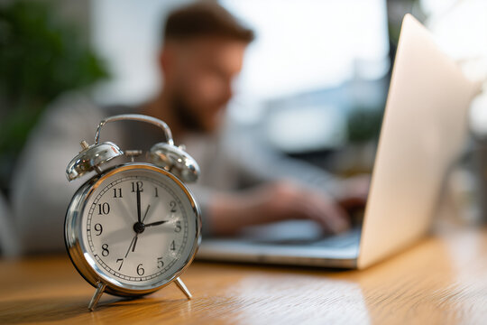 Selective focus of alarm clock on table near man using laptop at home, concept of time management