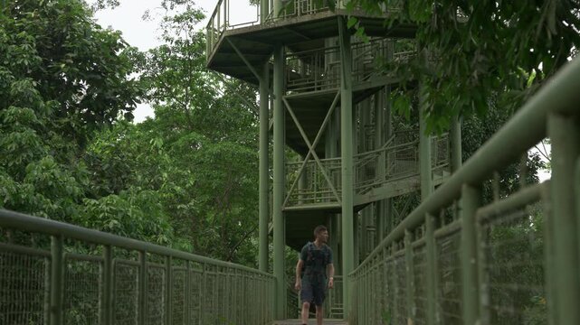 POV-style static shot on the RDC canopy walkway as a man approaches from the distance, walking toward the camera before passing out of frame. Tropical forest surrounds the elevated skywalk.