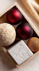 A close-up overhead view of a wooden tray containing decorative objects: two dark red matte spheres, a textured beige sphere, a white textured cube, and a golde