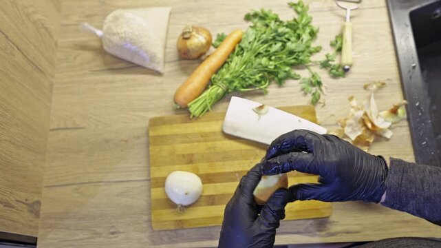 A shot of hands in black gloves meticulously peeling and slicing a fresh onion on a wooden chopping board. Fresh vegetables are visible, emphasizing healthy food preparation in a modern kitchen.