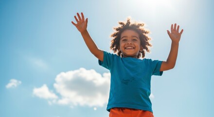Happy young boy with curly hair raising arms up against bright blue sky