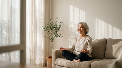 Senior asian woman meditating peacefully on a cozy sofa in a sunlit living room at home, finding inner peace