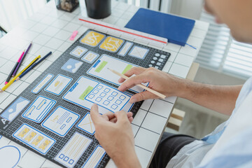 A designer is developing UX UI layouts, reviewing mobile app prototypes, and arranging interface elements on paper and board. He is focused on user experience and digital creativity.