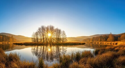 Fototapeta premium Sunrise over a calm lake with a small island of trees reflecting in the water, surrounded by golden autumn reeds and rolling hills under a clear blue sky