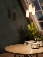 Coffee cup and books on round wooden table with hanging lamp under roof window in dim lit attic room
