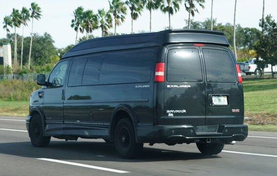 Tampa, Florida, U.S - Nov 10, 2025 - A dark-colored GMC Savana 2500 Explorer conversion van traveling on a highway flanked by palm trees.